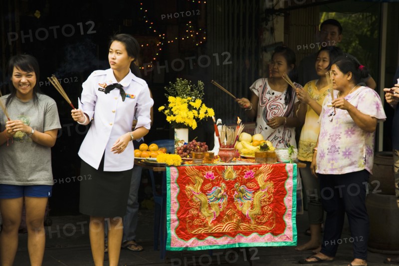 Woman in military uniform holding joss sticks beside offerings on decorated table celebrating local temple parade.