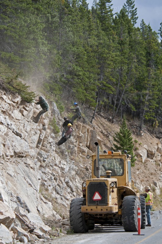 Workers wearing safety helmets on ropes clearing a rock slide and making the rock face safe along the Akamina Parkway Caterpillar tractor on parkway