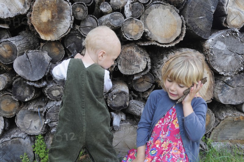 5 year old Eva and Oscar aged two playing beside a log pile with Eva using bark as pretend mobile phone.