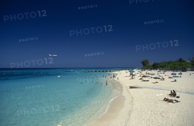 West Indies, Jamaica, Montego Bay, Sandy beach beside aquamarine water with people sunbathing and in the sea with low flying aeroplane over the water. Photo : David Cumming