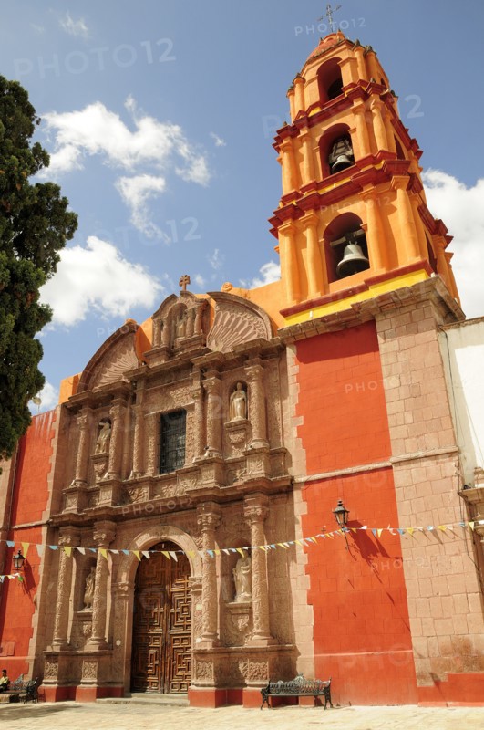 Mexico, Bajio, San Miguel de Allende, Exterior facade and bell tower of orange and yellow painted church building with flags hanging across doorway.. Photo : Nick Bonetti