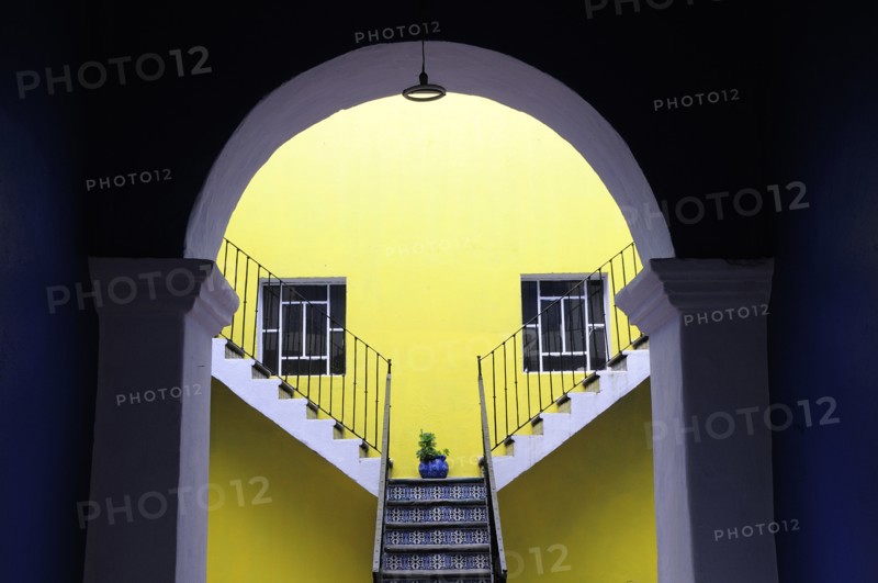 Mexico, Puebla, Looking into inner courtyard through archway in shadow towards yellow painted wall with blue and white tiled steps plant in blue pot and pair of square recessed windows. Photo : Nick Bonetti