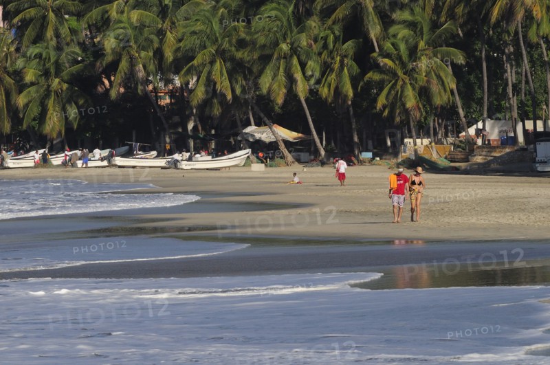 Mexico, Oaxaca, Puerto Escondido, Playa Marinera with couple walking along shore line of boats pulled up onto sand and overhanging palms beyond. Photo : Nick Bonetti