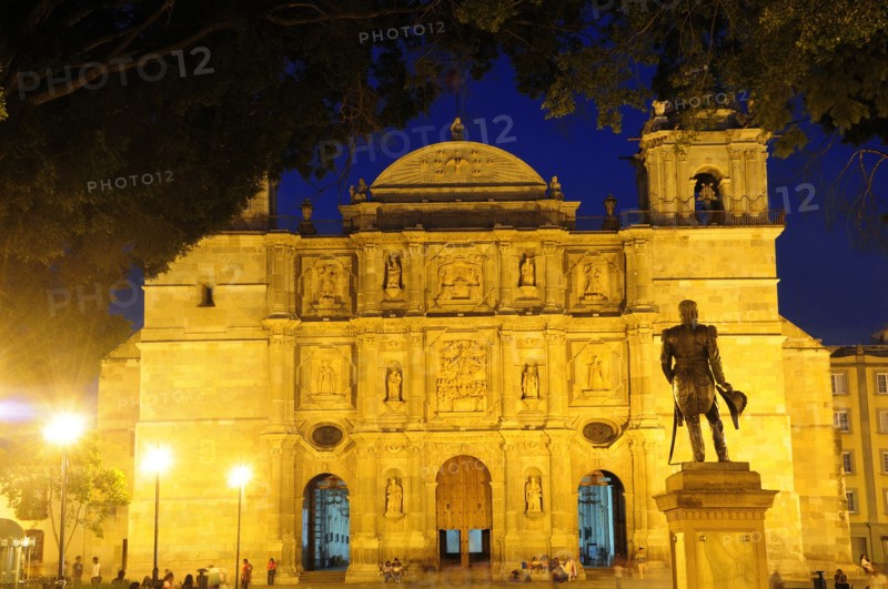 Mexico, Oaxaca, Baroque exterior facade of cathedral at night part framed by tree branches with people and statue in foreground. Photo : Nick Bonetti