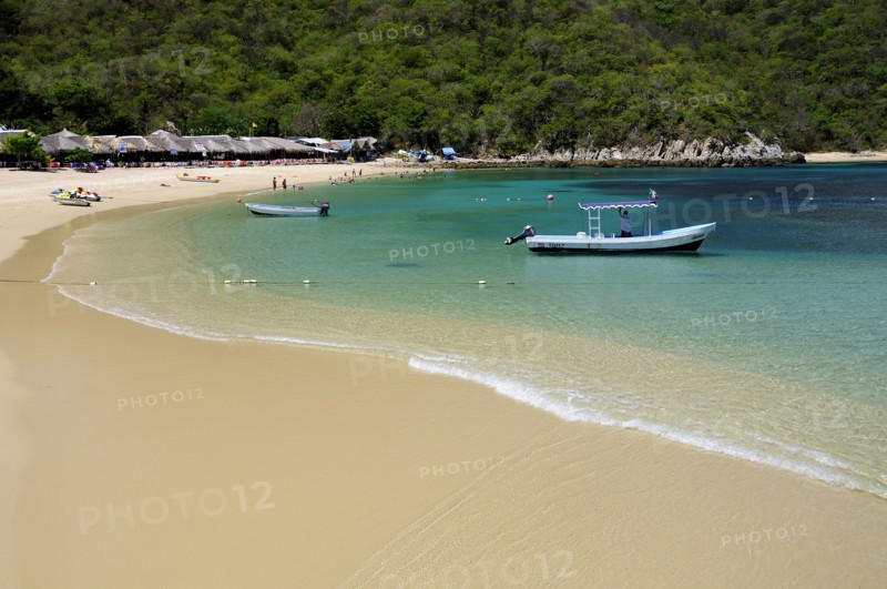 Mexico, Oaxaca, Huatulco, Playa La Entrega Stretch of sandy beach lined with thatched open fronted restaurants with tour boat in shallow water in foreground. Photo : Nick Bonetti