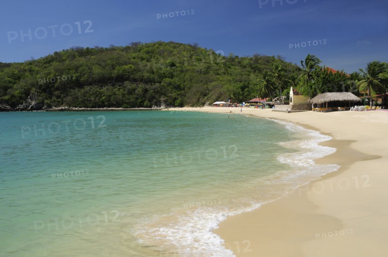 Mexico, Oaxaca, Huatulco, Sandy beach at Bahia Santa Cruz lined by restaurants and other buildings with tree covered coastline beyond. Photo : Nick Bonetti