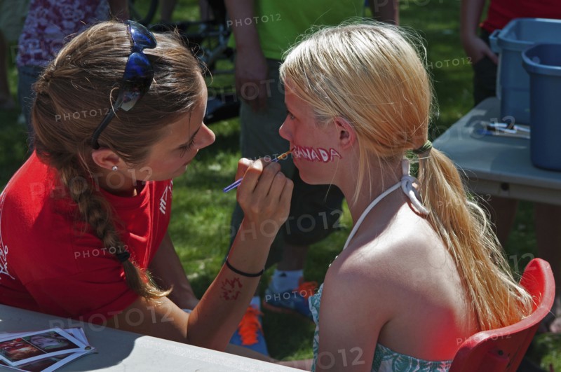 Canada, Alberta, Lethbridge, A bare-shouldered young blonde Canadian girl sits to have the word CANADA painted on her face at Canada Day celebrations at Henderson Park, Face painter in a red T shirt has a maple leaf, Canada's national emblem, stencilled on her forearm. 
Photo Trevor Page