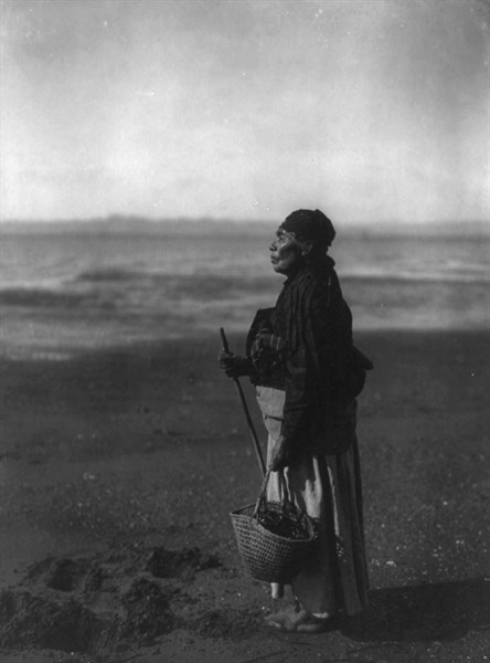 American Indian woman standing holding basket on beach