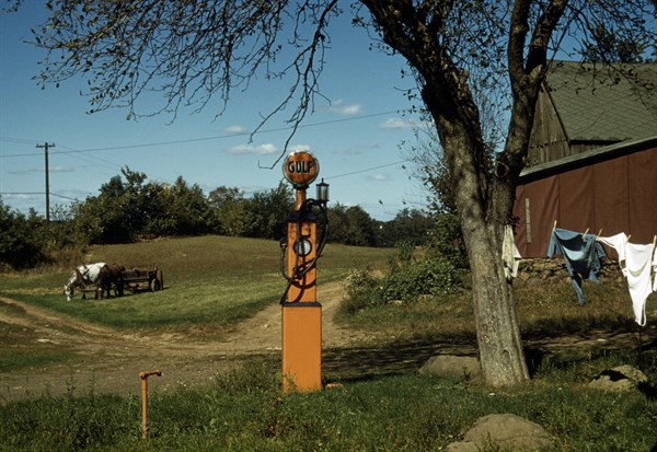Gas pump with clothesline, barn and horse-drawn wagon in background.