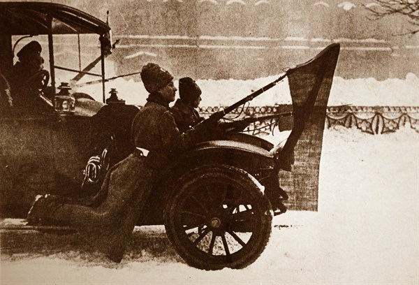 Photograph of soldiers riding on the mud flaps of a car with red flags attached to their guns in Petrograd