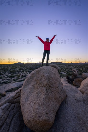 Young woman standing with outstretched arms on round granite rocks at sunset