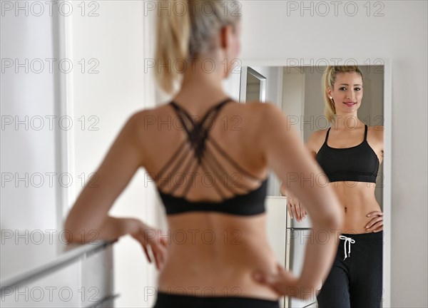 Young woman with sports bra and sweatpants looks at herself in the mirror