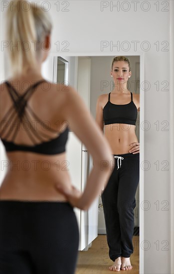 Young woman with sports bra and sweatpants looks at herself in the mirror