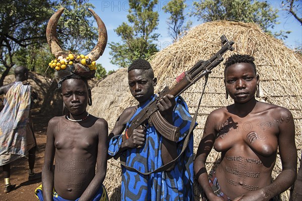 Young girls with decorative scars and man with Kalashnikov in front of hut