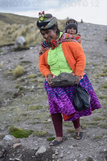 Young woman with child in a sling carrying a heavy stone to secure the shores of an artificial lake for irrigation