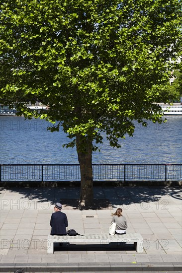 Two people sitting on concrete bench by river with tree