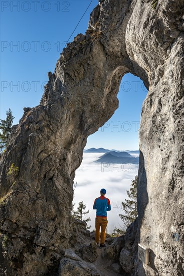 Young man stands in the rock gate Breitensteinfensterl with a view of the high fog above the valley