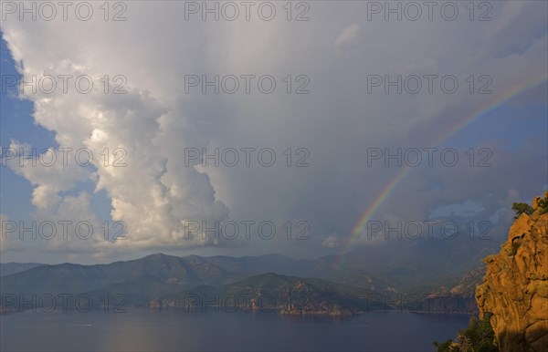 The typical bizarre red rocks of the Calanche of Piana and the mediterranean sea at the Gulf of Porto in the background below a rainbow and some clouds. The Calanche of Piana is in the western part of...