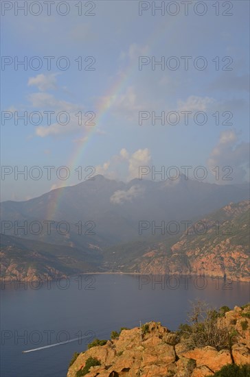 The typical bizarre red rocks of the Calanche of Piana and the mediterranean sea at the Gulf of Porto in the background below a rainbow and some clouds. The Calanche of Piana is in the western part of...