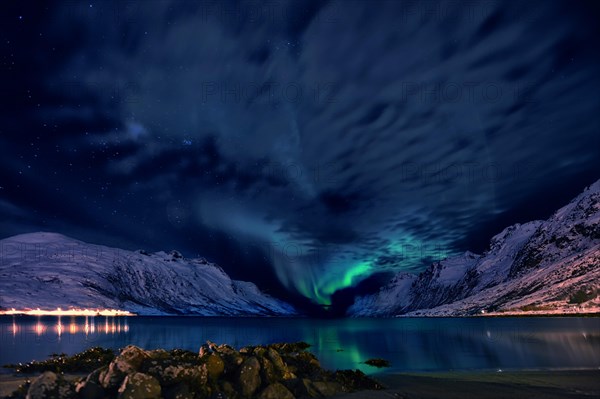 Aurora over a fjord with snow-covered mountains and an illuminated village