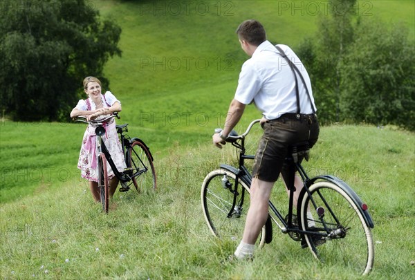 Man wearing leather pants and a woman wearing a dirndl on old bicycles within a natural landscape