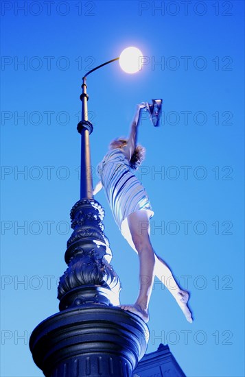 Woman wearing a summer dress standing on street lamp and holding hershoes in the air