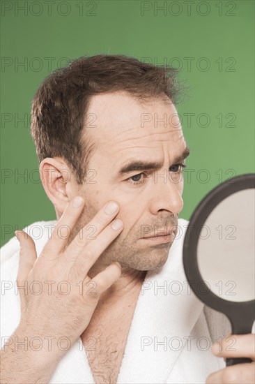 Man wearing a bathrobe observing himself critically in a hand mirror