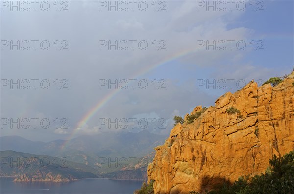 The typical bizarre red rocks of the Calanche of Piana and the mediterranean sea at the Gulf of Porto in the background below a rainbow and some clouds. The Calanche of Piana is in the western part of...