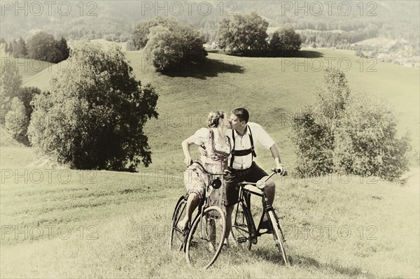 Man wearing leather pants and a woman wearing a dirndl kissing on old bicycles within a natural landscape