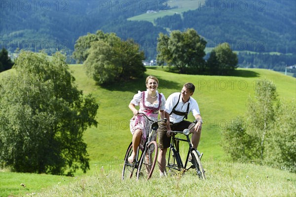 Man wearing leather pants and a woman wearing a dirndl on old bicycles within a natural landscape