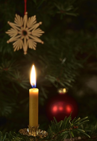 Burning candle in front of a Christmas tree with a star made of straw and a Christmas bauble
