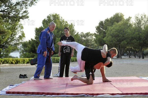 Two men doing martial arts on a mat in a park being watched by a trainer and his student