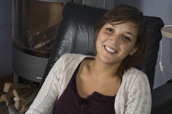 A young woman of Indonesian origin is sitting on a leather armchair in front of a wood burner