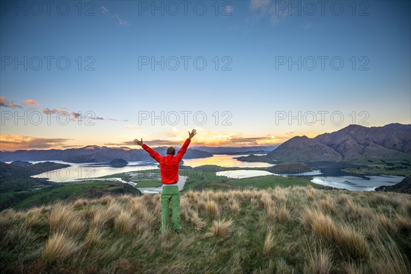 Hiker stretches his arms in the air, panoramic view of Wanaka Lake and mountains at sunset