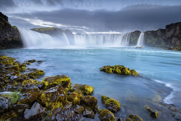 Long time exposure of a turquoise waterfall in volcanic landscape with dramatic clouds and green moss on rocks