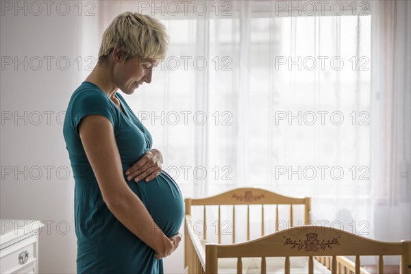 Pregnant woman holding her belly by baby cot next to the window