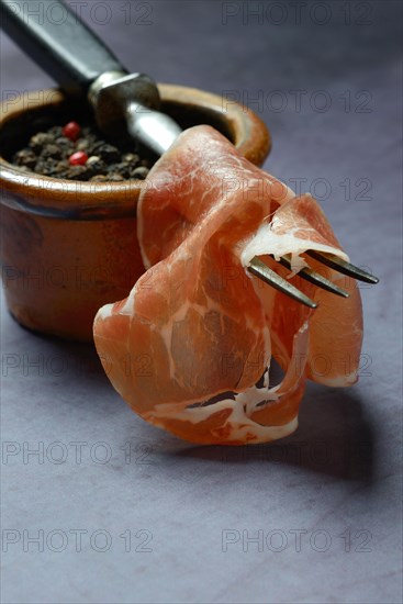 A slice of coppa on fork and clay pot with peppercorns