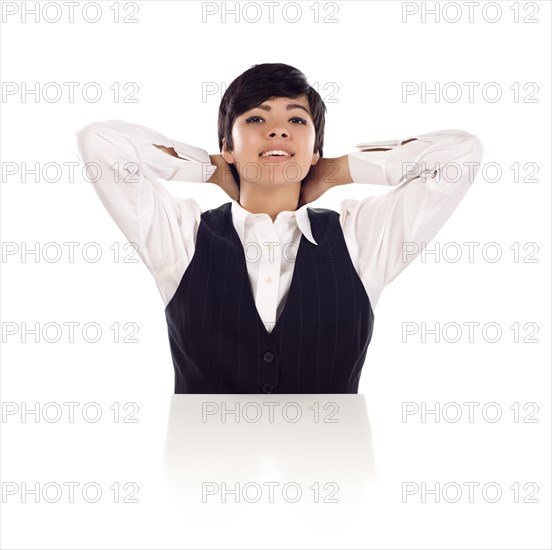Attractive smiling mixed-race young adult female sitting at white table with hands behind her head isolated on a white background