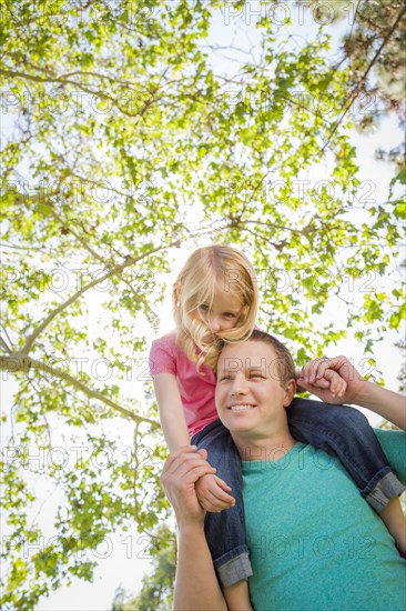 Cute young girl rides piggyback on her dads shoulders outside at the park