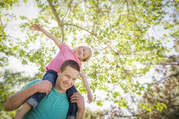 Cute young girl rides piggyback on her dads shoulders outside at the park