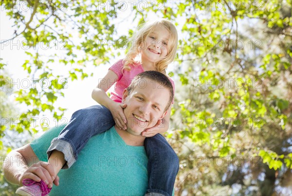 Cute young girl rides piggyback on her dads shoulders outside at the park