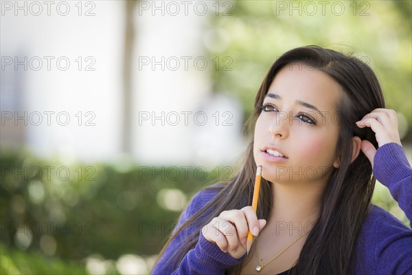 Attractive pensive mixed-race female student with pencil sitting on campus bench
