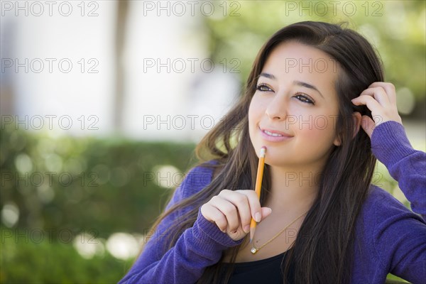 Attractive pensive mixed-race female student with pencil sitting on campus bench