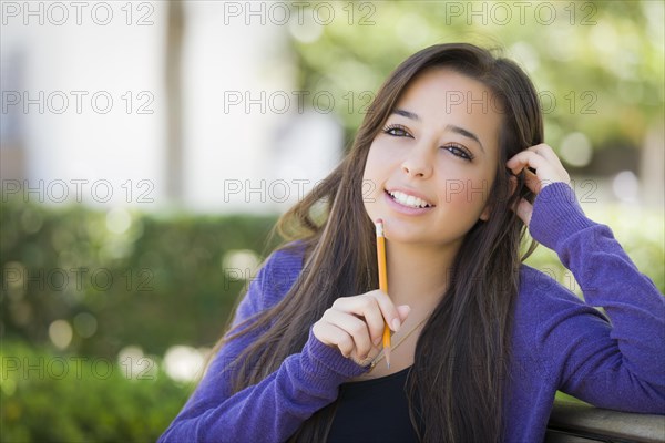 Attractive pensive mixed-race female student with pencil sitting on campus bench