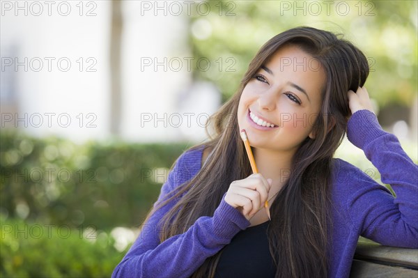 Attractive pensive mixed-race female student with pencil sitting on campus bench