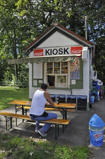 Man in white vest and jeans sits on bench in front of kiosk