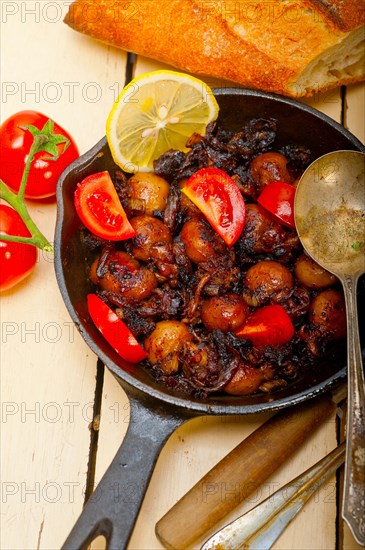 Baby cuttle fish roasted on iron skillet with tomatoes and onions over rustic wood table