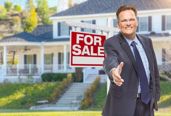 Smiling male agent reaching for a hand shake in front of beautiful house and for sale real estate sign