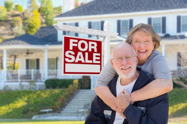 Senior adult couple in front of home for sale real estate sign and beautiful house