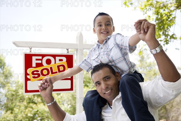 Excited hispanic father and son in front of sold for sale real estate sign